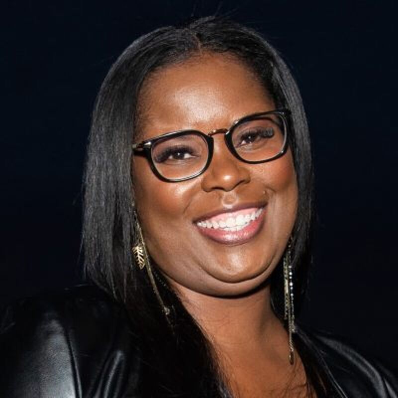 The image shows a woman with dark hair and glasses. She is smiling and appears to be in a well-lit environment. She is wearing a black jacket and earrings. The background is dark, which makes her the focal point of the image.